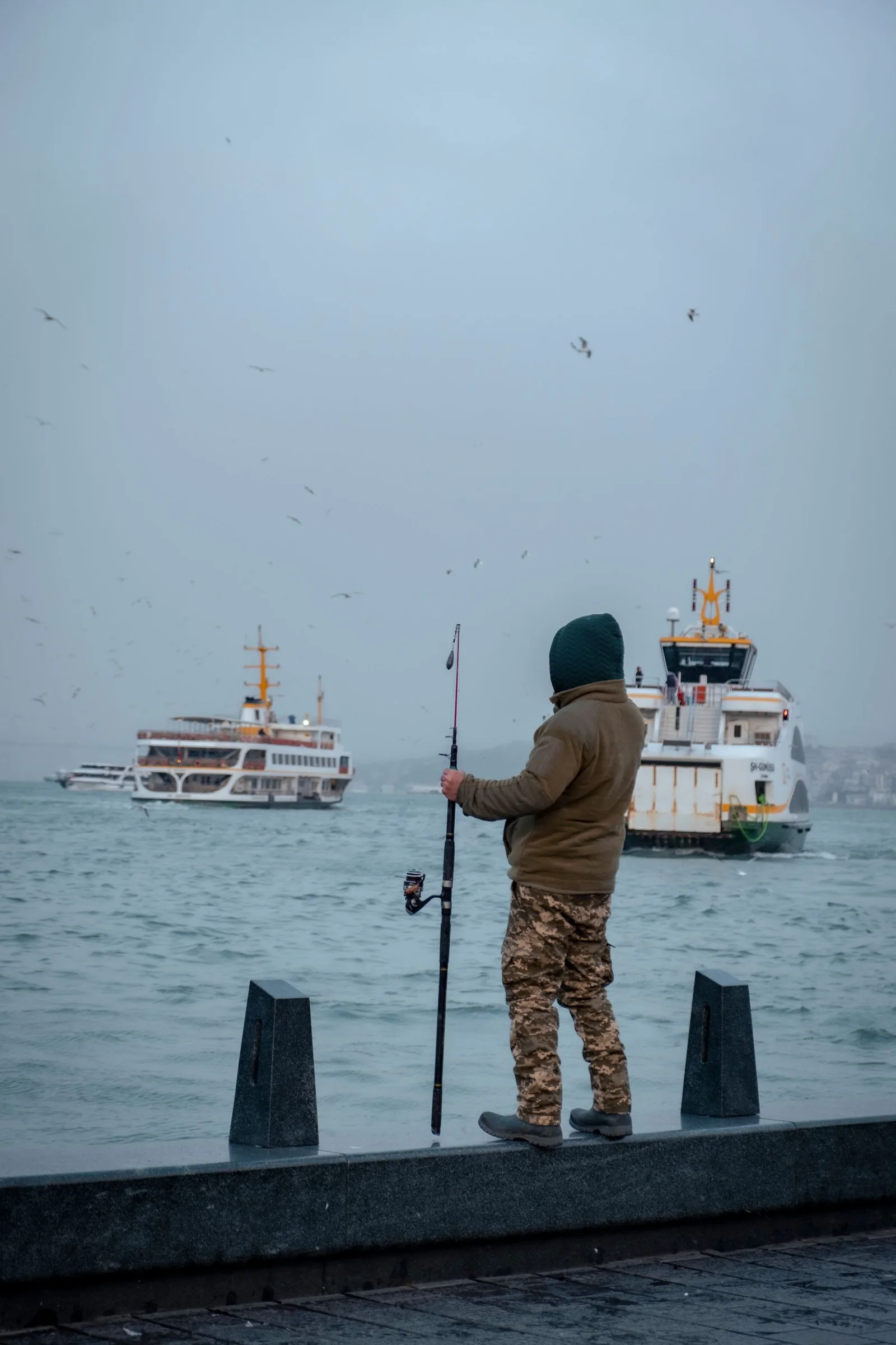 Man holding a fishing rod beside boats on an overcast shore.